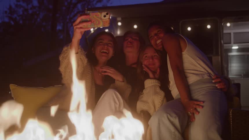 One woman takes a selfie with three other women sitting around a fire pit