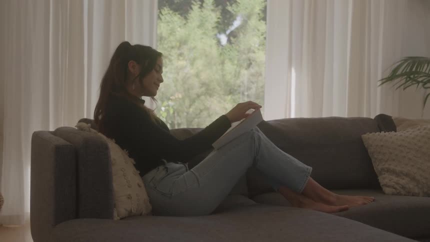 Young woman sitting on a couch with her feet up reading a book at midday
