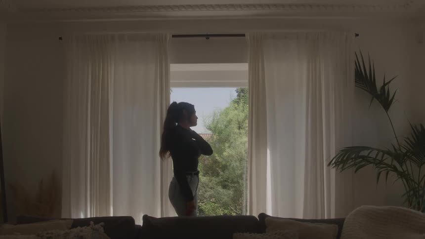 Young woman walking around the living room at midday looking bored and tired positioned just behind a couch