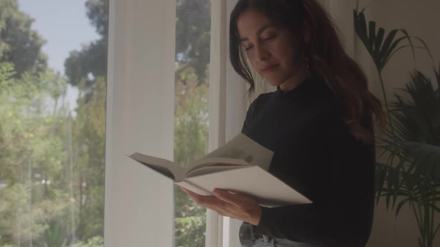 Woman standing reading a book next to the window