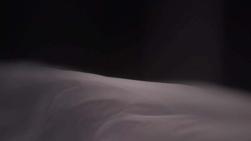 A close view of a persons body covered with a thin gray sheet as it slowly shifts creating gentle wrinkles and folds