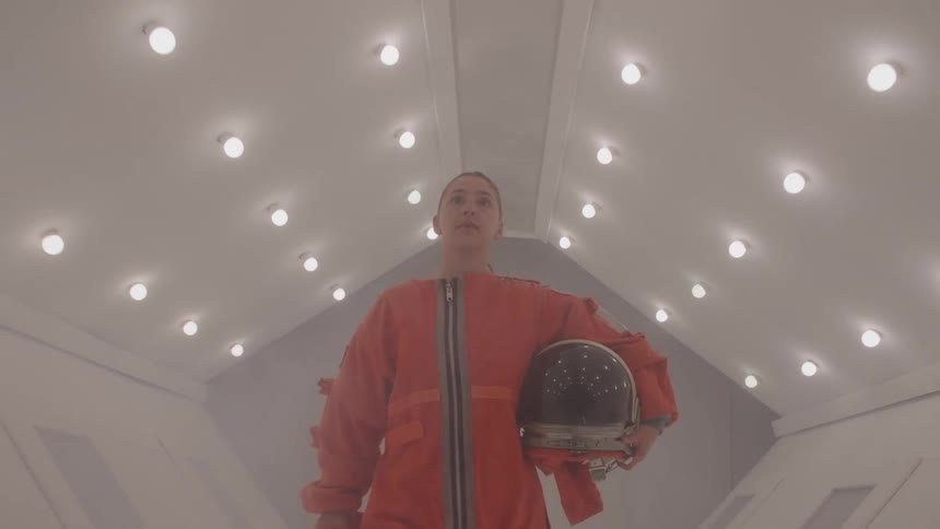 A woman in an orange space suit walks toward the camera down a sterile white corridor while holding a helmet under her arm