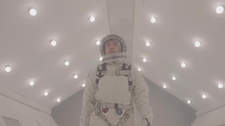 A low angle view of a man in a white and gray space suit walking down a white sterile corridor with many circular lights on the ceiling