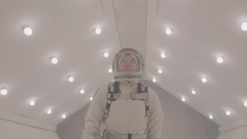 A woman in an astronaut suit stands in a white room with lights on the ceiling she is looking at the camera with a serious and determined expression on her face