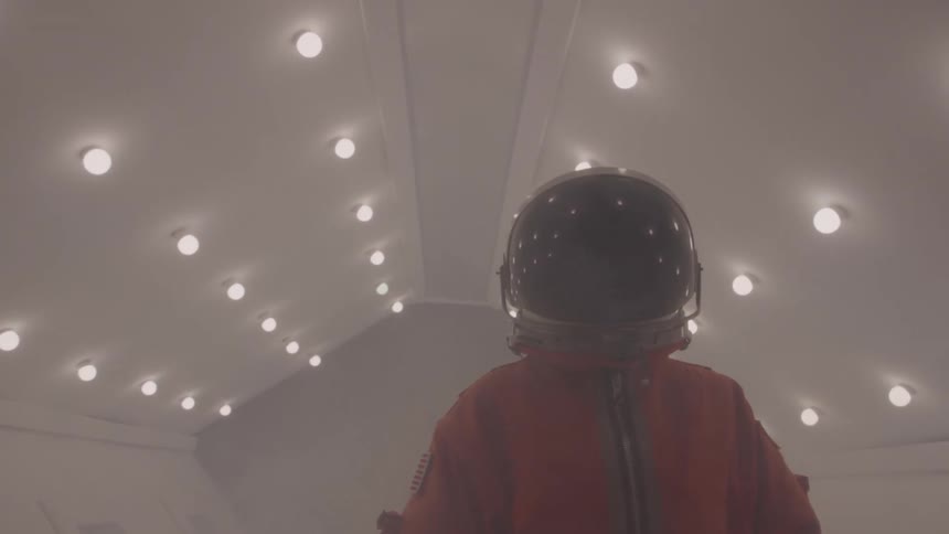 A person in a bulky orange space suit turns around in a hazy white corridor with many circular lights on the ceiling