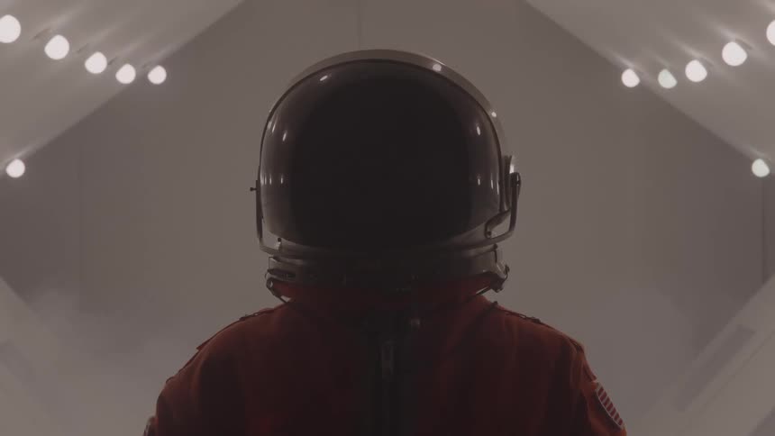 A person wearing a spacesuit and a helmet is standing in a brightly lit futuristic corridor the lighting creates a strong contrast silhouetting the person as they look straight ahead