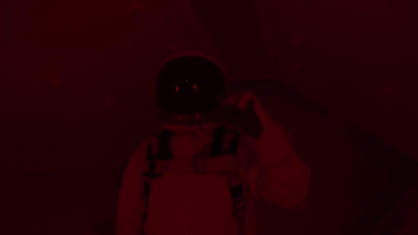 A man in a white space suit stands holding a helmet in his hand while a red and blue light fills the dark sterile room