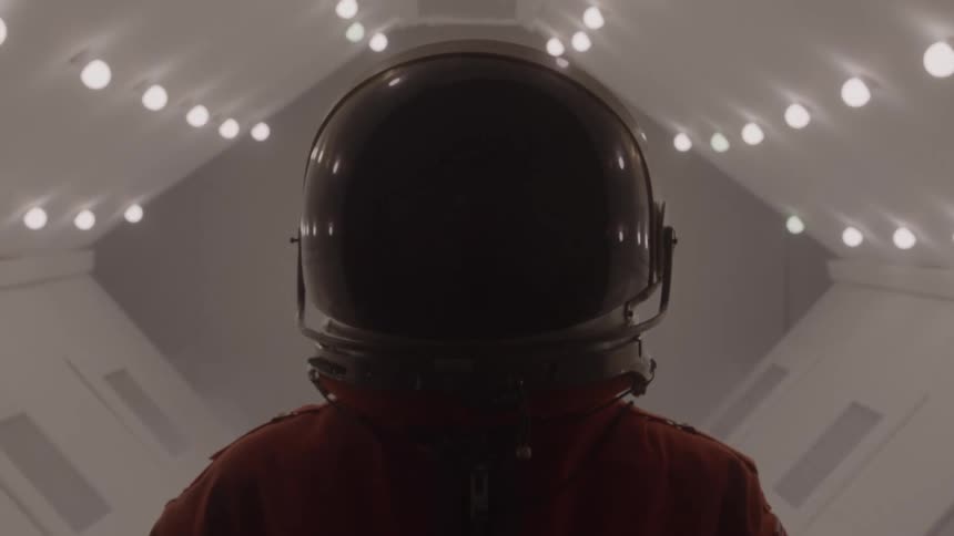 Person wearing a helmet and orange spacesuit inside a spaceship illuminated by a series of small lights