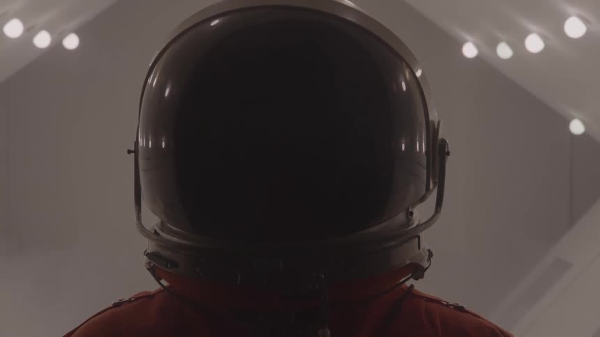 Person wearing a spacesuit with a dark reflective helmet in a room lit by multiple ceiling lights