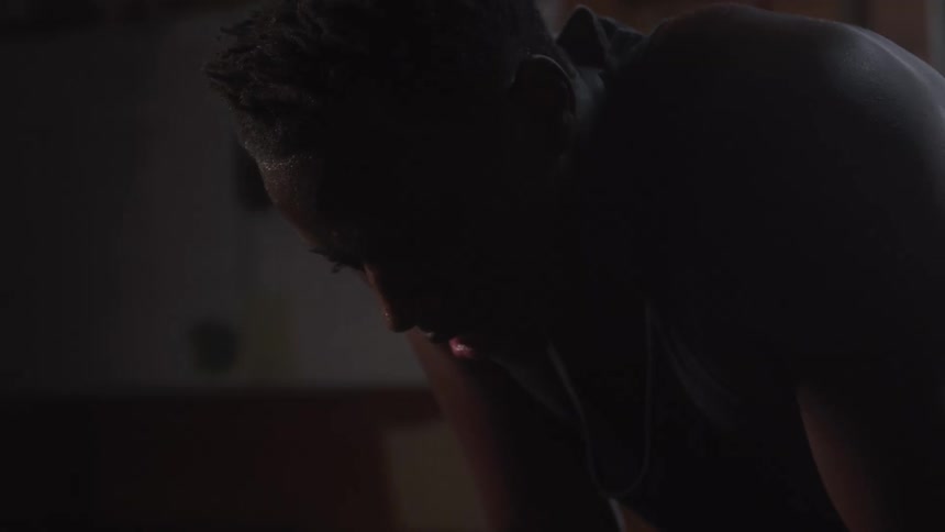 A close view of a man in a black tank top bending over with his hands on his knees and breathing heavily after a workout in a dark gym