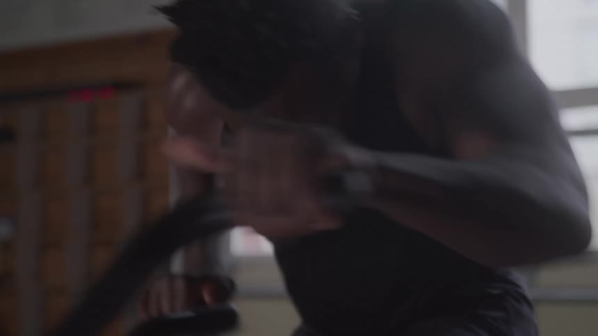 A man wearing a black tank top and shorts pedals an exercise bike with intense effort in a dimly lit gym with large window