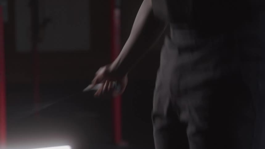 A man in a dark tank top skips rope vigorously his arm muscles tense with exertion in the low light