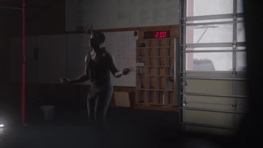 A woman is jumping rope in a gym with a large timer visible on the wall behind her the woman is wearing dark workout clothes and is focused on her exercise