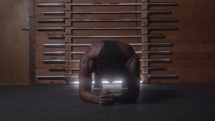 A man wearing a black tank top is holding a plank on a black rubber floor in a dimly lit gym with metal barbells on a wooden wall in the background