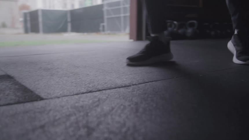 A close view of a mans legs and black sneakers as he stands on a rubber floor practicing box with a green field kettlebells and a building visible through an open door