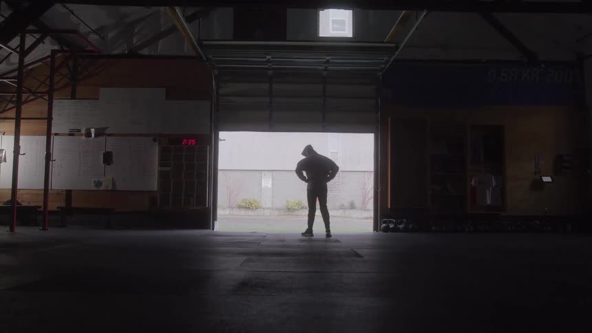 A silhouetted man wearing a hoodie and shorts stands in the center of a large empty gym with a bright garage door open behind him