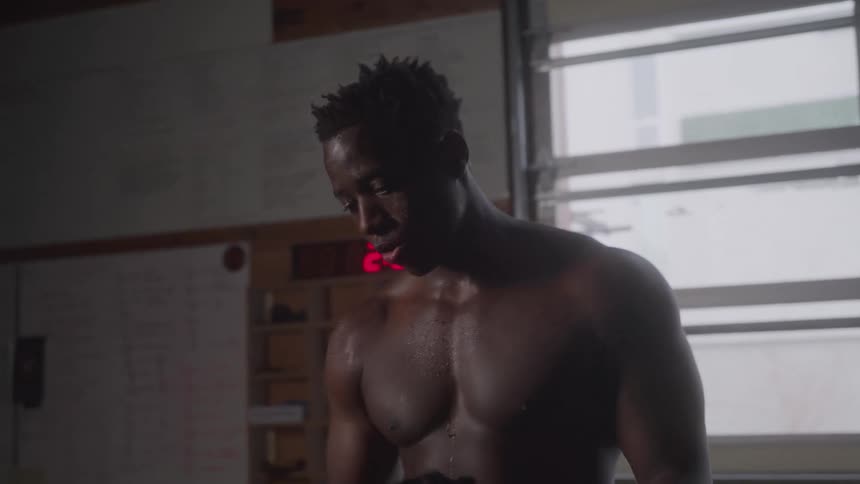 A man takes off his black tank top and stands shirtless with his hands on his knees breathing heavily after a workout in a dim gym