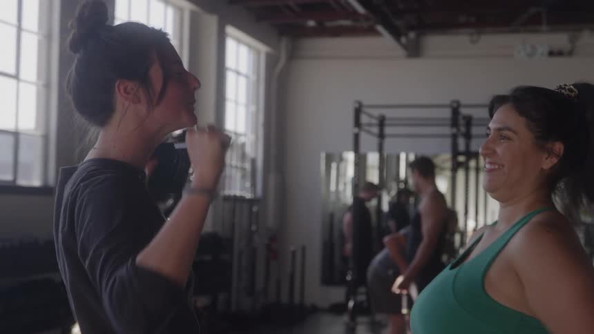 The video shows a medium shot of two women talking and laughing in a gym setting one woman is looking and smiling at the other who is explaining something with her hands