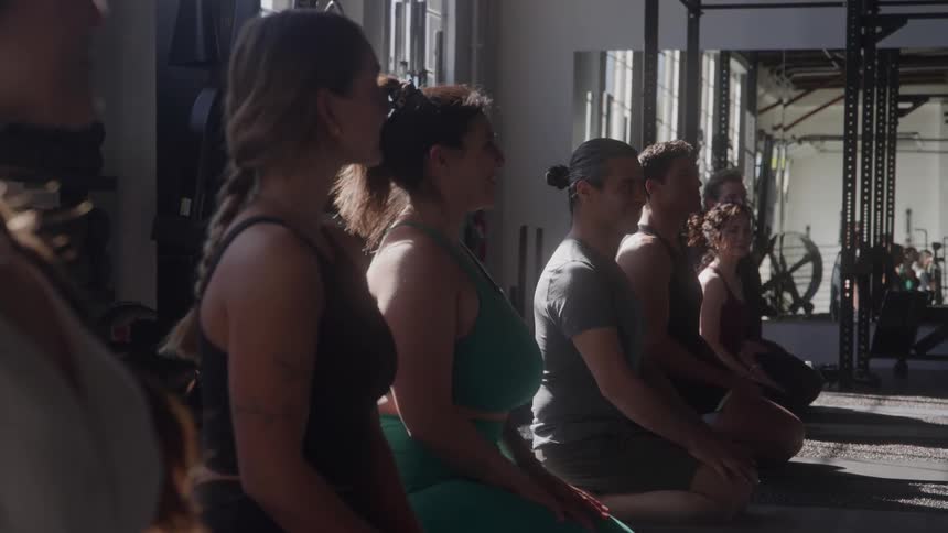 A diverse group of people is sitting in a row on the floor doing yoga they are all facing forward with their hands together in a prayer position and their eyes closed in concentration