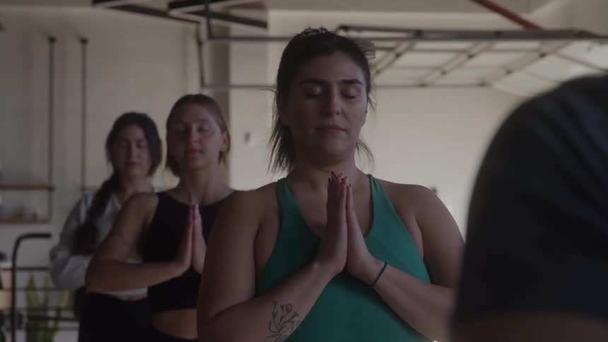 A yoga instructor leads a small class of three women with the instructor in the foreground and the students behind her they are all standing with their eyes closed and their hands in a prayer position