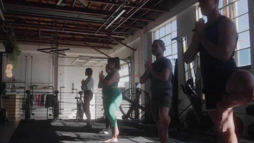 A midwide shot shows a group of people doing yoga in a gym with exposed ceilings and large windows the people are in a tree pose with their hands together in front of their chests