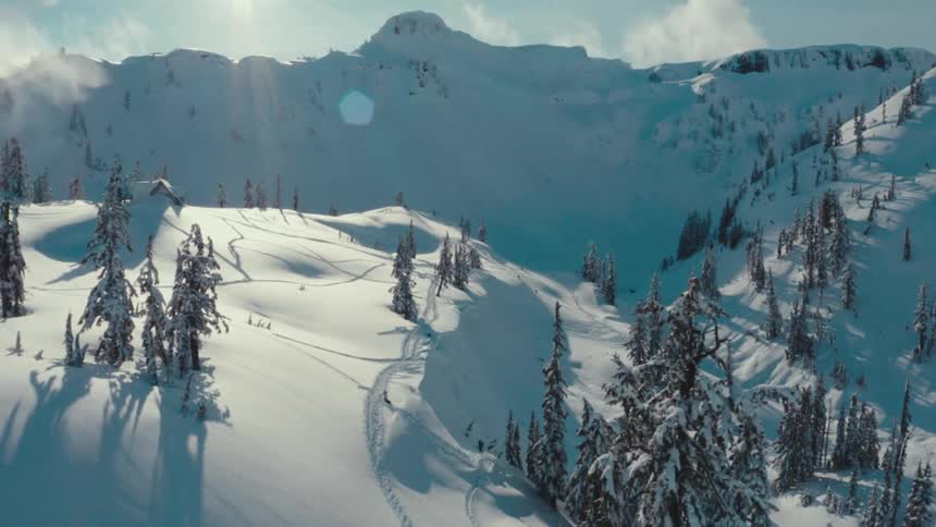 A drone shot shows a beautiful snowy mountain landscape with deep snow tall pine trees and tracks from skiers or snowboarders