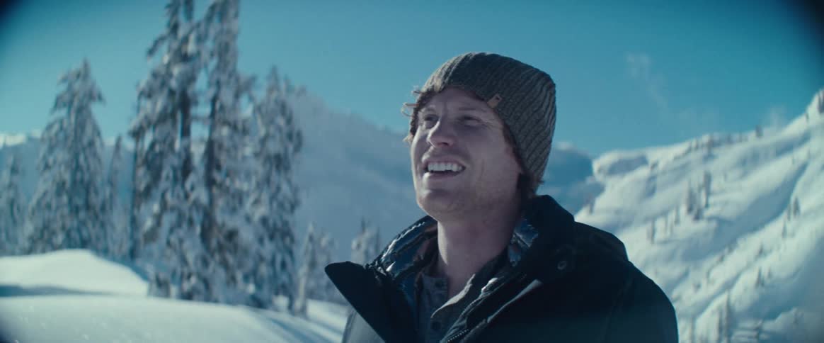 A close view of a man with a beard and a knit cap smiling while looking at a snowy landscape with pine trees and a bright clear sky