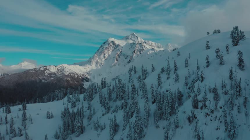Forest occupies the narrow valley between a forested snowy mountain and a tall craggy mountain on a cloudy winter day