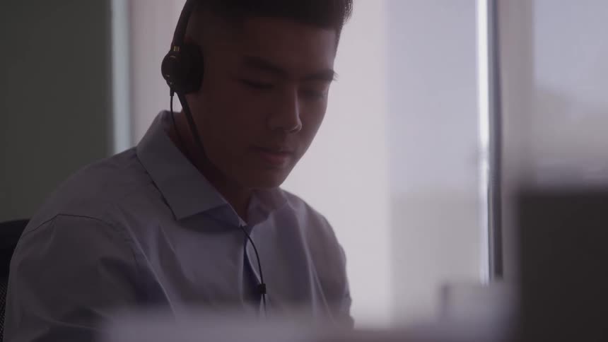 The video shows a man working at a computer in a lowlit office he is wearing a headset and appears to be concentrating intently on his work suggesting he is on a call or in a virtual meeting