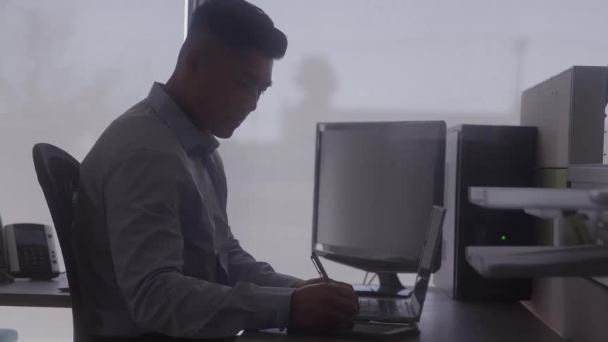 A man works at his office desk on a laptop with a monitor nearby