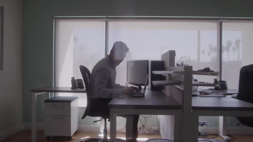 A man wearing a light blue dress shirt sits at a desk in a bright office with large windows and white blinds working on a laptop with a monitor next to it