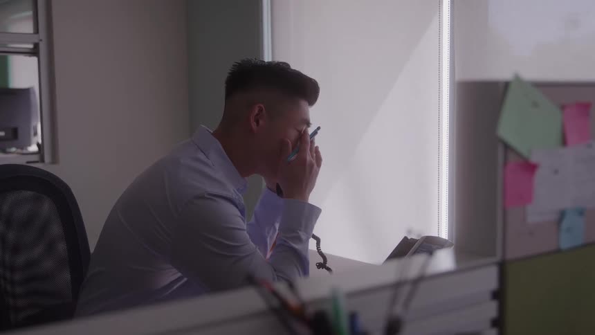 A male professional is sitting at a desk in an office facing away from the camera while working on a laptop the scene is brightly lit from the windows creating a silhouette effect that focuses on the individual and his task