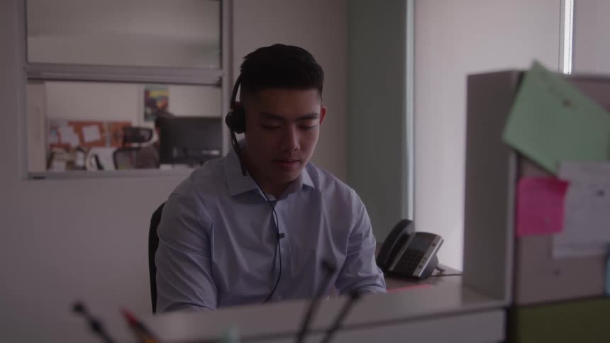 A young man sits at a desk in an office and is wearing a headset and answering a call