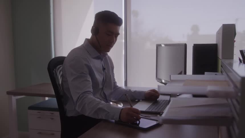 A person sits at a desk in an office using a pen on a tablet while wearing a headset and looking at a computer
