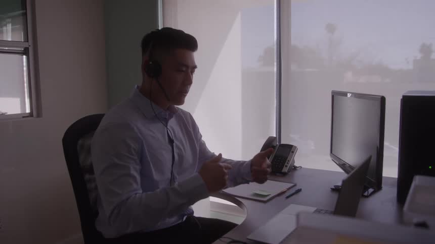 A man wearing a headset sits at a desk with a computer and phone actively engaged in a conversation and gesturing with his hands