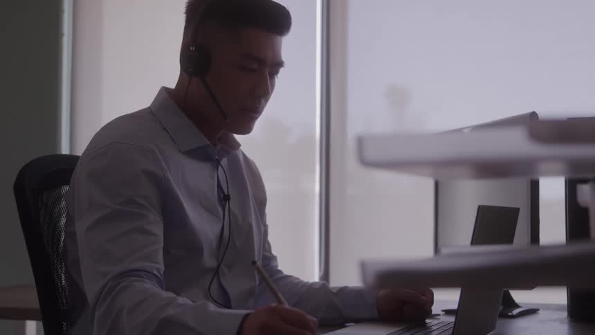 A man wearing a headset sits at a desk in front of a window focused on writing notes while participating in a call