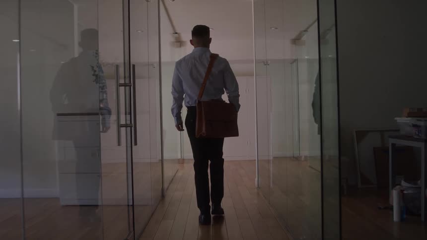 A man wearing a dress shirt and jeans with a messenger bag walks down a long office hallway lined with glass doors with reflections and bright lighting