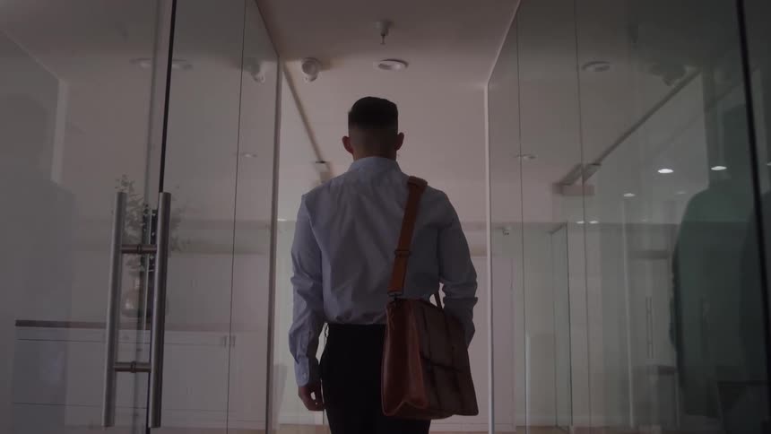 A man with a shoulder bag walks through a glass walled office hallway