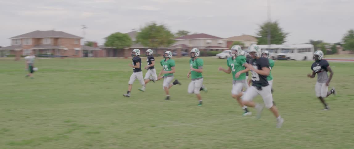 A group of young football players in green and black jerseys are jogging on a grassy field they are wearing helmets and a few of them are carrying footballs