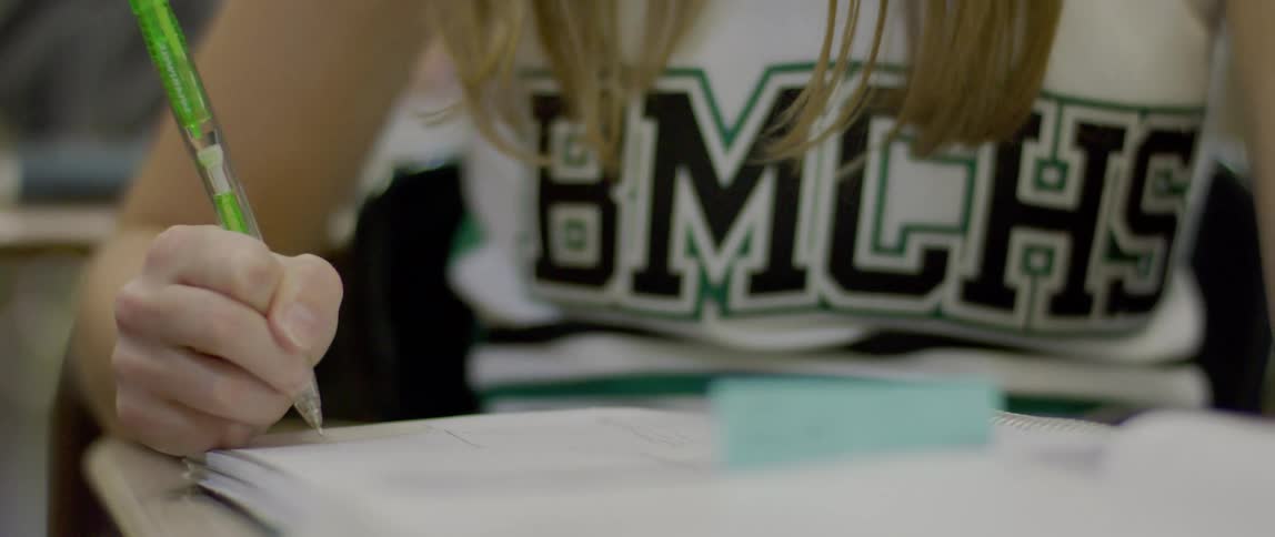 Female student inside a classroom wearing a cheerleader jersey writing in a notebook with a pen