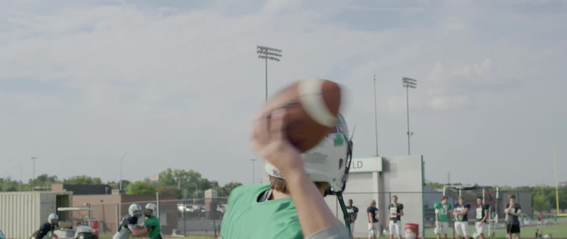 A football player wearing a green jersey with the number 12 and a white helmet with a green stripe on top throws a football the players last name mcguinness is written on the back of his helmet