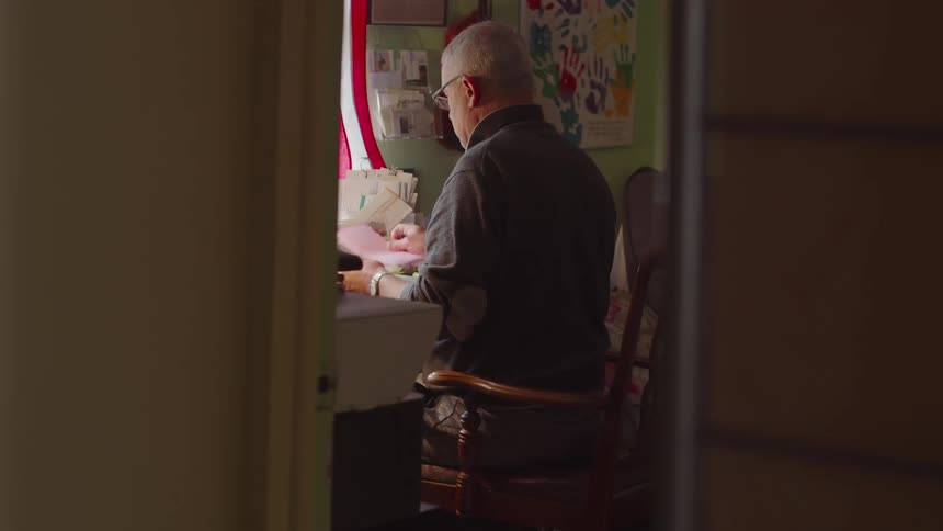An elderly man sitting on a wooden chair and examining papers in an indoor setting