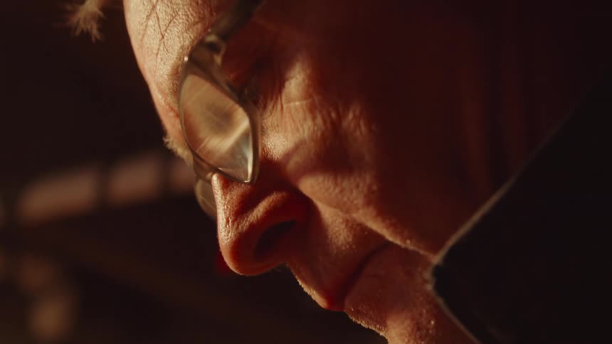 A close side view of a man with wrinkles and glasses looking down at something in a dimly lit space