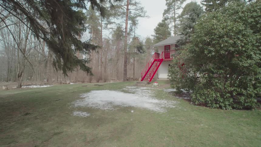The video shows a wide shot of a large twostory house with a red staircase on the side the house is surrounded by tall bare trees and patches of melting snow on the ground indicating its early spring