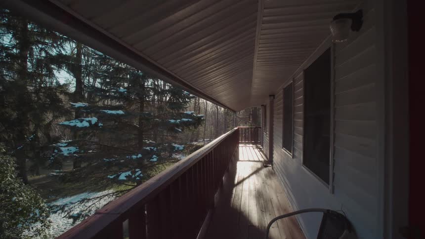 A slow moving shot travels down the long sunlit porch of a building overlooking a snowy wooded area