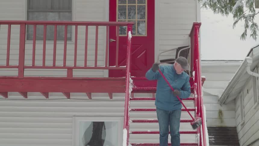 A man in a blue jacket and cap carefully clears snow from bright red outdoor stairs in front of a white house working with steady focus