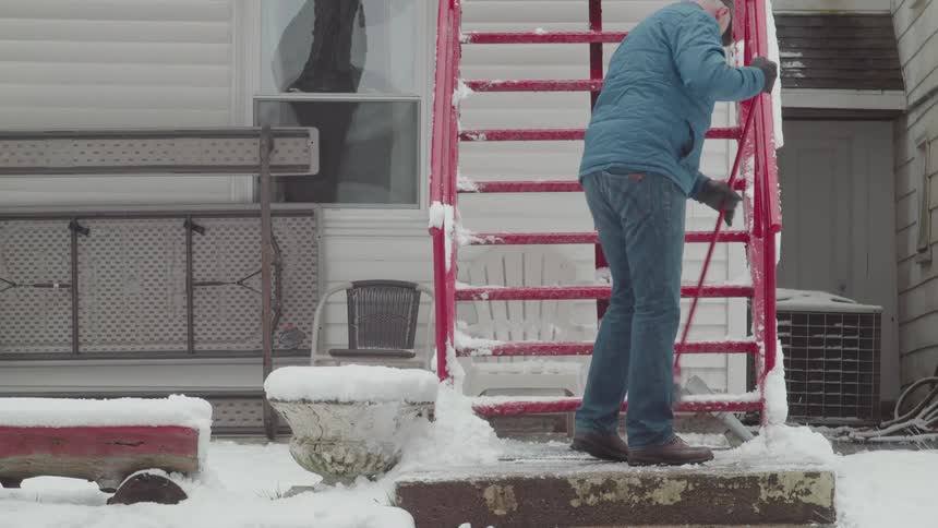 A wide shot of a snowy house with red stairs shows a man in a blue jacket using a broom to clear the snow from the steps he sweeps a pile of snow off the bottom step