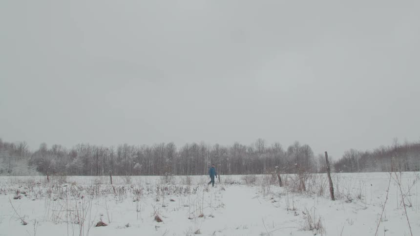 Adult man wearing a baseball cap and a blue jacket walking on the snow next to a fence with a forest on the background outdoors during day time