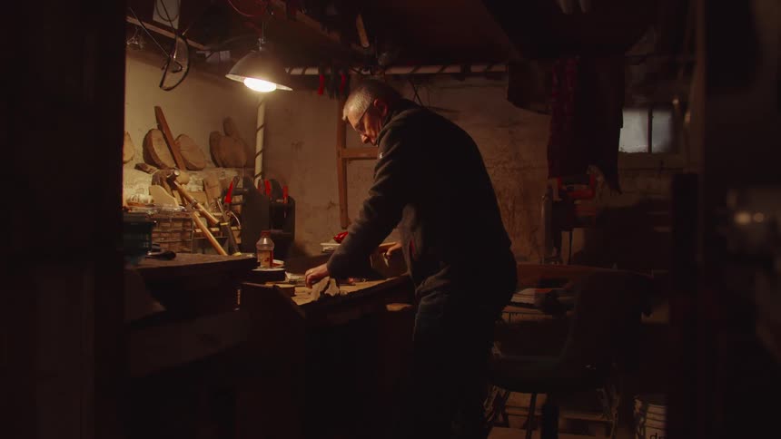 A man works at a cluttered workbench in a dimly lit rustic workshop