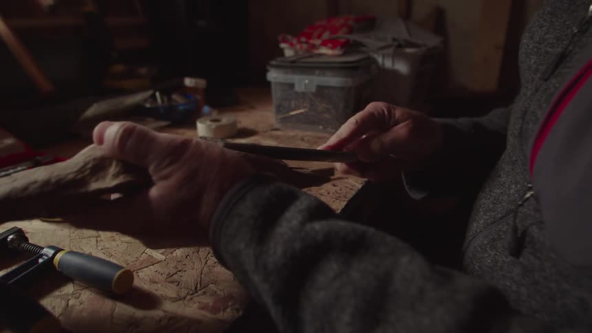 In a dimly lit workshop a person carefully files a piece of wood at a cluttered workbench